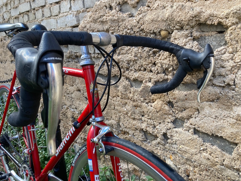 Vélo de course Bianchi de couleur rouge le long d'un mur. Vue trois quart avant avec zoom sur détails du cintre, notamment les cocottes Campagnolo Mirage Avanti